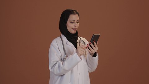 استخدام الأجهزة التقنية الحديثة ودمج التكنولوجيا مع الطب, تقديم الخدمات والرعاية الصحية عبر الإنترنت، بورتريه مقرب لطبيبة عربيه خليجيه سعودية ترتدي المعطف الطبي و السماعه تقوم باستخدام الهاتف المحمول، التطبيقات والبرامج الصحية، خلفية بنية
