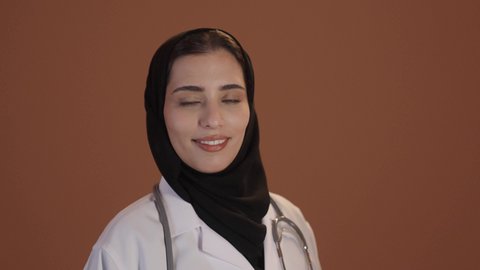 الوقوف بايدى متقاطعة، بورتريه لطبيبة عربية خليجية سعودية ترتدي العباءة والحجاب مع المعطف الابيض تقف باستقامة، النظر بايماءات السعادة والسرور، مهن ووظائف نسائية، تقديم الخدمات للمرضى بكل كفاءه، خلفيه بنية