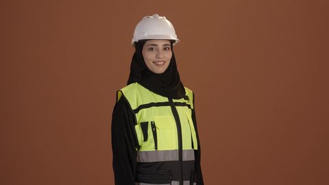 تحقيق حلم النجاح والوصول لمهنة الهندسة بتعابير الفخر، المشاريع الهندسية ومخططات الإعمار, بورتريه مقرب لفتاة عربية خليجية سعودية مبتسمة ترتدي سترة الحماية والخوذة وتنظر إلى الكاميرا بايماءات السعادة والسرور, العمل في القطاع الهندسي، خلفية بنية