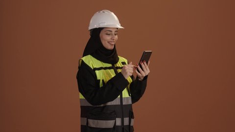 Working in the engineering sector, women's professions and jobs, portrait of a Saudi Arabian Gulf female engineer wearing a hijab, black abaya, helmet, and protective vest, holding a mobile phone, using technological devices, gestures of happiness and pride, brown background