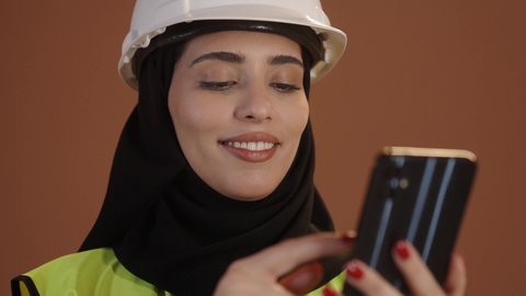 استخدام الأجهزة التقنية الحديثة, بورتريه لمهندسة عربية خليجية سعودية ترتدي الحجاب والعباءة السوداء والخوذة وسترة الحماية تحمل الهاتف المحمول، ايماءات السعادة والفخر، مهن ووظائف نسائية ، العمل في القطاع الهندسي, خلفية بنية