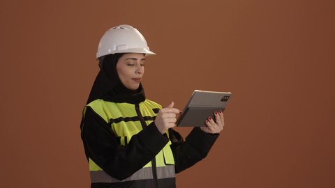 Technological development and progress in the field of industry and engineering; monitoring and supervising work via tablet; portrait of a Saudi Arabian Gulf female engineer wearing a hijab, black abaya, and protective vest, holding a tablet in her hand; brown background.