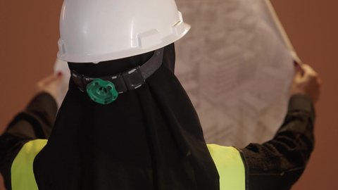 Development and progress in the field of engineering, architectural design and plan review, close-up portrait from behind of a veiled Saudi Arabian Gulf female engineer wearing a protective vest and helmet, holding a construction plan, engineering project management, women's professions and jobs, brown background
