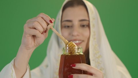 بورتريه لإمرأة عربية خليجية سعودية ترتدي الثوب الابيض المطرز بالذهبي وتضع الشيله على رأسها تحمل بيدها وعاء زجاجي ملئ بالعسل ، ارتداء الزي السعودي التقليدي الشعبي, استخراج عسل النحل الطبيعي من المناحل، منتج غذائي محلي وطني في المملكة العربية السعودية، الأغذية الصحية المفيدة، خلفية خضراء, كروما