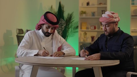 Completing office tasks, writing and recording notes, understanding and cooperating among colleagues to achieve the company's goals, verifying and auditing financial offers, developing a plan and work mechanism, an Arab Gulf Saudi man wearing traditional attire and a ghutrah explaining tasks to his colleague.