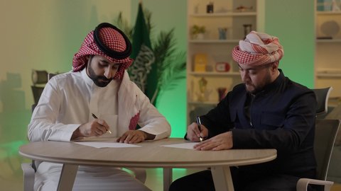 كتابة وتسجيل الملاحظات, إنجاز الأعمال المكتبية، التفاهم والتعاون بين الزملاء لتحقيق أهداف الشركة، التحقق والتدقيق في العروض المالية، تطوير خطة وآلية العمل، رجل عربي خليجي سعودي يرتدي الزي التقليدي والشماغ يقوم بشرح المهام لزميله