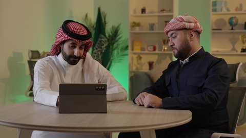 Gestures of integration and focus, dialogue and discussion among colleagues, using a tablet to accomplish projects and tasks, spending enjoyable time with friends, an Arab Gulf Saudi man wearing traditional attire and a ghutrah sitting with his friend at work, integrating advanced technology in the workplace.