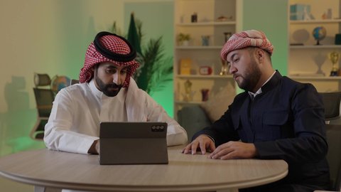 رجل عربي خليجي سعودي يرتدي الزى التقليدي والشماغ  يجلس مع صديقه في العمل ، ايماءات الاندماج والتركيز ، الحوار والنقاش بين زملاء العمل، استخدم جهاز التابلت لانجاز المشاريع والمهام ، قضاء وقت ممتع مع الأصدقاء, دمج التكنولوجيا المتطورة فى العمل