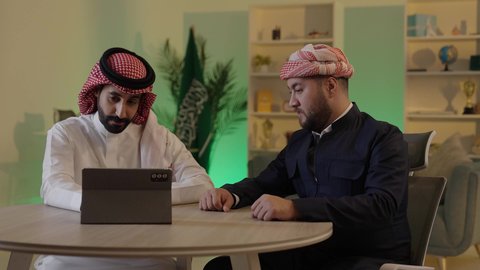 قضاء وقت ممتع مع الأصدقاء, ايماءات الاندماج والتركيز ، الحوار والنقاش بين زملاء العمل، استخدم جهاز التابلت لانجاز المشاريع والمهام ،رجل عربي خليجي سعودي يرتدي الزى التقليدي والشماغ  يجلس مع صديقه في العمل ، دمج التكنولوجيا المتطورة فى العمل