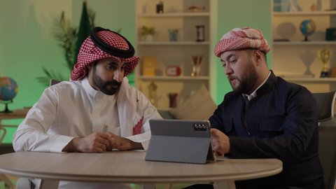 الحوار والنقاش بين زملاء العمل، ايماءات الاندماج والتركيز ،استخدم جهاز التابلت لانجاز المشاريع والمهام ، قضاء وقت ممتع مع الأصدقاء, رجل عربي خليجي سعودي يرتدي الزى التقليدي والشماغ  يجلس مع صديقه في العمل ، دمج التكنولوجيا المتطورة فى العمل