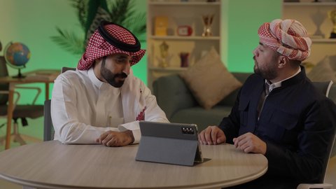استخدم جهاز التابلت لانجاز المشاريع والمهام ، قضاء وقت ممتع مع الأصدقاء, رجل عربي خليجي سعودي يرتدي الزى التقليدي والشماغ  يجلس مع صديقه في العمل ، ايماءات الاندماج والتركيز ، الحوار والنقاش بين زملاء العمل ، دمج التكنولوجيا المتطورة فى العمل