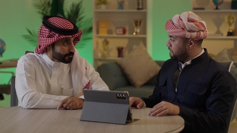 Gestures of integration and focus, dialogue and discussion among colleagues, using a tablet to accomplish projects and tasks, spending enjoyable time with friends, an Arab Gulf Saudi man wearing traditional attire and a ghutrah sitting with his friend at work, integrating advanced technology in the workplace.