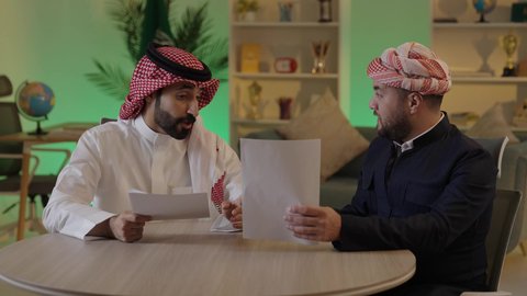 كتابة وتسجيل الملاحظات, إنجاز الأعمال المكتبية، التفاهم والتعاون بين الزملاء لتحقيق أهداف الشركة، التحقق والتدقيق في العروض المالية، تطوير خطة وآلية العمل، رجل عربي خليجي سعودي يرتدي الزي التقليدي والشماغ يقوم بشرح المهام لزميله