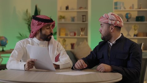 Completing office tasks, writing and recording notes, understanding and cooperating among colleagues to achieve the company's goals, verifying and auditing financial offers, developing a plan and work mechanism, an Arab Gulf Saudi man wearing traditional attire and a ghutrah explaining tasks to his colleague.