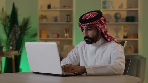 Managing and organizing business affairs through technical devices, conducting remote business meetings, integrating work with technology and tech, browsing social media, an Arab Gulf Saudi businessman wearing traditional attire and a ghutrah working on a laptop with focused gestures.