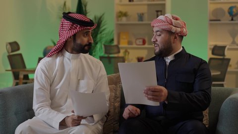Completing office tasks, writing and recording notes, understanding and cooperating among colleagues to achieve the company's goals, verifying and auditing financial offers, developing a plan and work mechanism, an Arab Gulf Saudi man wearing traditional attire and a ghutrah explaining tasks to his colleague.