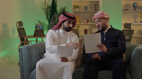 Writing and recording notes, completing office tasks, understanding and cooperating among colleagues to achieve the company's goals, verifying and auditing financial offers, developing a plan and work mechanism, an Arab Gulf Saudi man wearing traditional attire and a ghutrah explaining tasks to his colleague.