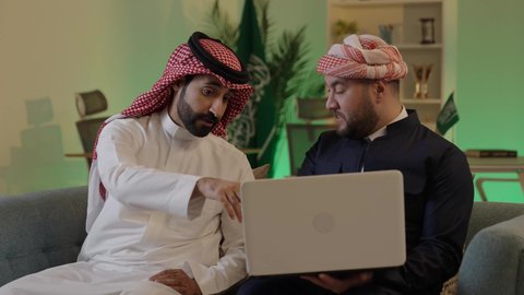 Gestures of integration and focus, dialogue and discussion among colleagues, using a laptop to accomplish projects and tasks, spending enjoyable time with friends, an Arab Gulf Saudi man wearing traditional attire and a ghutrah sitting with his friend at work, integrating advanced technology in the workplace.