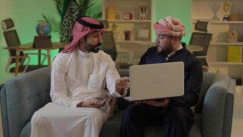 Spending quality time with friends, gestures of engagement and focus, dialogue and discussion among colleagues, using a laptop to accomplish projects and tasks, an Arab Gulf Saudi man wearing traditional attire and a ghutrah sitting with his friend at work, integrating advanced technology in the workplace.