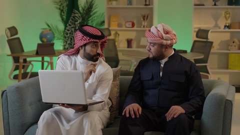 استخدم جهاز اللابتوب لانجاز المشاريع والمهام ، قضاء وقت ممتع مع الأصدقاء, رجل عربي خليجي سعودي يرتدي الزى التقليدي والشماغ  يجلس مع صديقه في العمل ، دمج التكنولوجيا المتطورة فى العمل، ايماءات الاندماج والتركيز ، الحوار والنقاش بين زملاء العمل