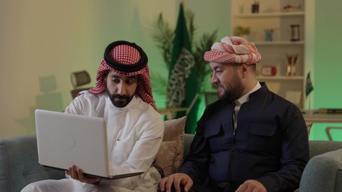 دمج التكنولوجيا المتطورة فى العمل، ايماءات الاندماج والتركيز ، الحوار والنقاش بين زملاء العمل، استخدم جهاز اللابتوب لانجاز المشاريع والمهام ، قضاء وقت ممتع مع الأصدقاء, رجل عربي خليجي سعودي يرتدي الزى التقليدي والشماغ  يجلس مع صديقه في العمل ، دمج التكنولوجيا المتطورة فى العمل