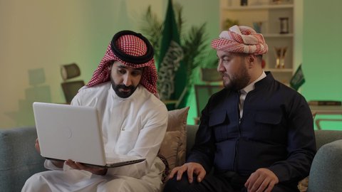 الحوار والنقاش بين زملاء العمل، ايماءات الاندماج والتركيز ، استخدم جهاز اللابتوب لانجاز المشاريع والمهام ، قضاء وقت ممتع مع الأصدقاء, رجل عربي خليجي سعودي يرتدي الزى التقليدي والشماغ  يجلس مع صديقه في العمل ، دمج التكنولوجيا المتطورة فى العمل