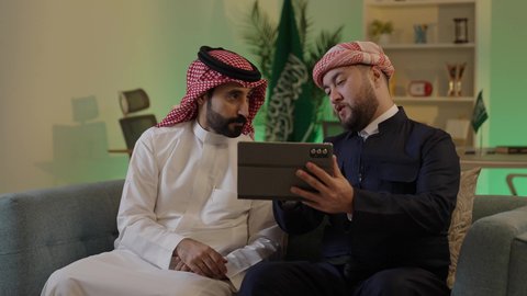 Gestures of integration and focus, dialogue and discussion among colleagues, using a tablet to accomplish projects and tasks, spending enjoyable time with friends, an Arab Gulf Saudi man wearing traditional attire and a ghutrah sitting with his friend at work, integrating advanced technology in the workplace.