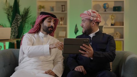 Gestures of integration and focus, dialogue and discussion among colleagues, using a tablet to accomplish projects and tasks, spending enjoyable time with friends, an Arab Gulf Saudi man wearing traditional attire and a ghutrah sitting with his friend at work, integrating advanced technology in the workplace.