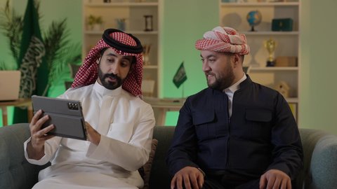 Spending quality time with friends, gestures of engagement and focus, dialogue and discussion among colleagues, using a tablet to accomplish projects and tasks, an Arab Gulf Saudi man wearing traditional attire and a ghutrah sitting with his friend at work, integrating advanced technology in the workplace.