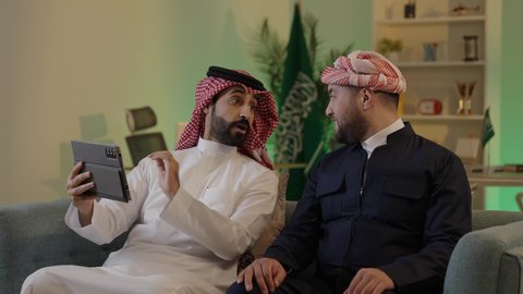 الحوار والنقاش بين زملاء العمل، ايماءات الاندماج والتركيز ، استخدم جهاز التابلت لانجاز المشاريع والمهام ، قضاء وقت ممتع مع الأصدقاء, رجل عربي خليجي سعودي يرتدي الزى التقليدي والشماغ  يجلس مع صديقه في العمل ، دمج التكنولوجيا المتطورة فى العمل