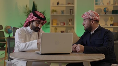 ايماءات الاندماج والتركيز ، الحوار والنقاش بين زملاء العمل، استخدم جهاز اللابتوب لانجاز المشاريع والمهام ، قضاء وقت ممتع مع الأصدقاء, رجل عربي خليجي سعودي يرتدي الزى التقليدي والشماغ  يجلس مع صديقه في العمل ، دمج التكنولوجيا المتطورة فى العمل