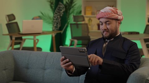 تصفح مواقع التواصل الاجتماعي، إدارة شؤون الأعمال وتنظيمها عبر الاجهزة التقنية، دمج العمل مع التقنية والتكنولوجيا،رجل اعمال عربي خليجي سعودي يرتدي الزي  الكاجوال والشماغ  يعمل على جهاز التابلت بإيماءات التركيز