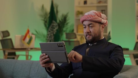 A Saudi Arabian Gulf businessman wearing casual attire and a shemagh is working on a tablet with focused gestures, managing and organizing business affairs through technological devices, integrating work with technology and tech, browsing social media.