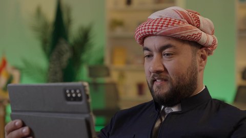 إدارة شؤون الأعمال وتنظيمها عبر الاجهزة التقنية، دمج العمل مع التقنية والتكنولوجيا، تصفح مواقع التواصل الاجتماعي، رجل اعمال عربي خليجي سعودي يرتدي الزي  الكاجوال والشماغ  يعمل على جهاز التابلت بإيماءات التركيز