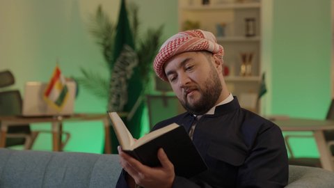 The importance of reading in expanding the horizons of knowledge and increasing information, an Arab Gulf Saudi man wearing casual attire and a shemagh sits on a comfortable sofa reading from a book, the role of education in developing Saudi society, knowledge is the key to development and success.
