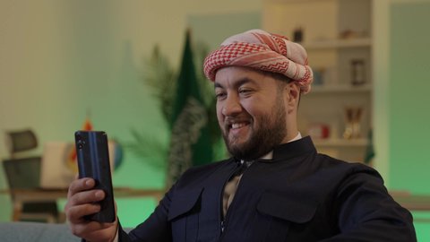 Using a modern technical device, an Arab Gulf Saudi man wearing casual attire and a shemagh sits in an office using a mobile phone with focused gestures, browsing social media, chatting with family and friends via mobile.