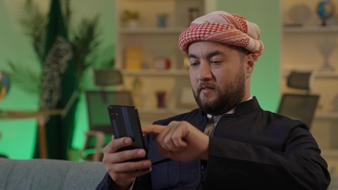 الدردشة مع الاهل والاصدقاء عبر الجوال، استخدام جهاز تقني حديث، رجل عربي خليجي سعودي يرتدي الزى الكاجوال والشماغ يجلس في مكتب يستخدام الهاتف المحمول بإيماءات التركيز، تصفح مواقع التواصل الاجتماعي