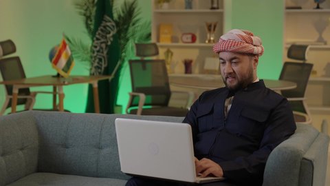 إدارة شؤون الأعمال وتنظيمها عبر الاجهزة التقنية، دمج العمل مع التقنية والتكنولوجيا، تصفح مواقع التواصل الاجتماعي، رجل اعمال عربي خليجي سعودي يرتدي الزي الكاجوال والشماغ  يعمل على جهاز الحاسوب المحمول بإيماءات التركيز