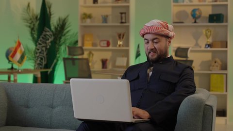 رجل اعمال عربي خليجي سعودي يرتدي الزي الكاجوال والشماغ  يعمل على جهاز الحاسوب المحمول بإيماءات التركيز، إدارة شؤون الأعمال وتنظيمها عبر الاجهزة التقنية، دمج العمل مع التقنية والتكنولوجيا، تصفح مواقع التواصل الاجتماعي