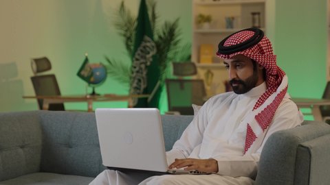 تصفح مواقع التواصل الاجتماعي، اجراء اجتماع عمل عن بعد ،  إدارة شؤون الأعمال وتنظيمها عبر الاجهزة التقنية، دمج العمل مع التقنية والتكنولوجيا، رجل اعمال عربي خليجي سعودي يرتدي الزي التقليدي والشماغ  يعمل على جهاز الحاسوب المحمول بإيماءات التركيز
