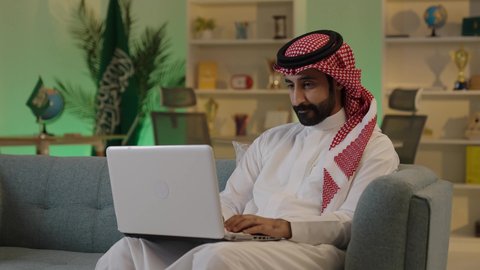 دمج العمل مع التقنية والتكنولوجيا، اجراء اجتماع عمل عن بعد ،  إدارة شؤون الأعمال وتنظيمها عبر الاجهزة التقنية،تصفح مواقع التواصل الاجتماعي، رجل اعمال عربي خليجي سعودي يرتدي الزي التقليدي والشماغ  يعمل على جهاز الحاسوب المحمول بإيماءات التركيز