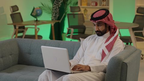 إدارة شؤون الأعمال وتنظيمها عبر الاجهزة التقنية، دمج العمل مع التقنية والتكنولوجيا، تصفح مواقع التواصل الاجتماعي، رجل اعمال عربي خليجي سعودي يرتدي الزي التقليدي والشماغ  يعمل على جهاز الحاسوب المحمول بإيماءات التركيز، اجراء اجتماع عمل عن بعد