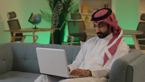 اجراء اجتماع عمل عن بعد ،  إدارة شؤون الأعمال وتنظيمها عبر الاجهزة التقنية، دمج العمل مع التقنية والتكنولوجيا، تصفح مواقع التواصل الاجتماعي، رجل اعمال عربي خليجي سعودي يرتدي الزي التقليدي والشماغ  يعمل على جهاز الحاسوب المحمول بإيماءات التركيز