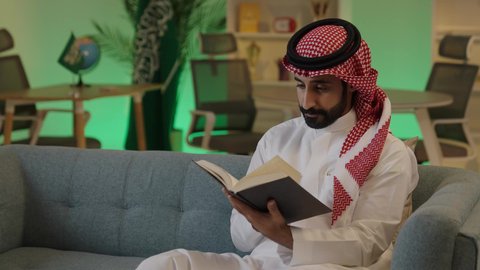 The importance of reading in expanding the horizons of knowledge and increasing information, an Arab Gulf Saudi man wearing traditional attire and a shemagh sits on a comfortable sofa reading from a book, the role of education in developing Saudi society, knowledge is the key to development and success.