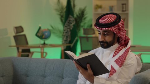 دور التعليم في تطوير المجتمع السعودي، أهمية القراءة في توسيع آفاق المعرفة وزيادة المعلومات، رجل عربي خليجي سعودي يرتدي الزي التقليدي والشماغ يجلس على اريكة مريحة يقرأ من كتاب، العلم مفتاح التطور والنجاح