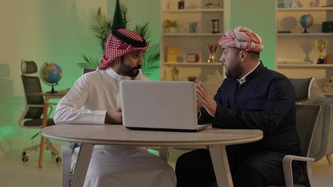 اجتماع في المكتب لمناقشة سير العمل, متابعة إستراتيجيات وأهداف الشركة, دراسة وتحليل المشاريع الجديدة, موظفان عربيان خليجيان سعوديان  يستخدمون جهاز اللابتوب المحمول، استخدام التقنيات الحديثة، مناقشة ومتابعة الاعمال، التعاون والتفاهم بين الموظفين، مهن ووظائف مكتبية