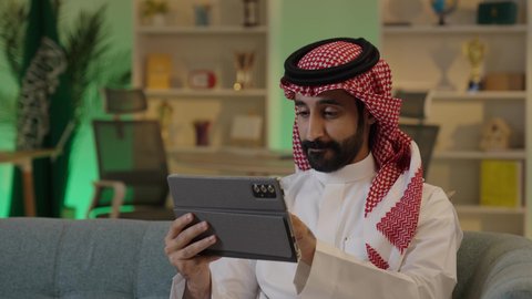 رجل اعمال عربي خليجي سعودي يرتدي الزي التقليدي والشماغ  يعمل على جهاز التابلت بإيماءات التركيز، إدارة شؤون الأعمال وتنظيمها عبر الاجهزة التقنية، دمج العمل مع التقنية والتكنولوجيا، تصفح مواقع التواصل الاجتماعي