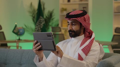 دمج العمل مع التقنية والتكنولوجيا، إدارة شؤون الأعمال وتنظيمها عبر الاجهزة التقنية، تصفح مواقع التواصل الاجتماعي، رجل اعمال عربي خليجي سعودي يرتدي الزي التقليدي والشماغ  يعمل على جهاز التابلت بإيماءات التركيز