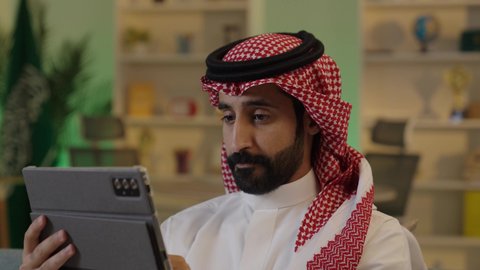 Managing and organizing business affairs through technical devices, integrating work with technology and tech, browsing social media, an Arab Gulf Saudi businessman wearing traditional attire and a ghutrah working on a tablet with focused gestures.