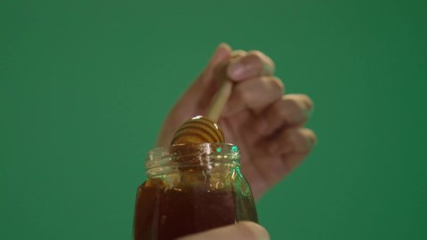 Extracting natural honey from beehives, local national food products in the Kingdom of Saudi Arabia, wearing traditional Saudi folk attire, healthy beneficial foods, a portrait of an Arab Gulf Saudi woman wearing a white dress embroidered with gold and wearing a shayla on her head, holding a glass container filled with honey in her hand, with a green background.